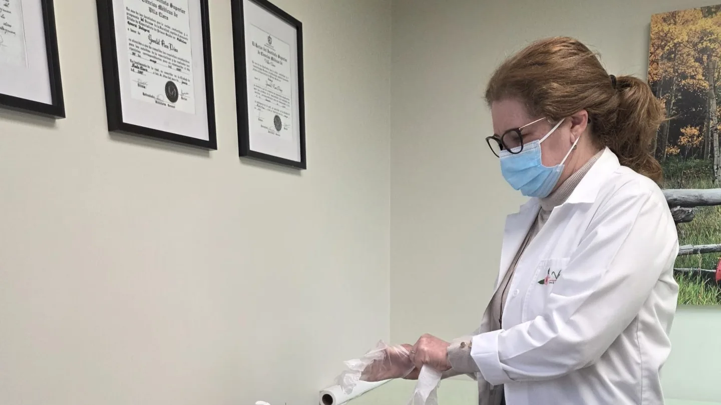 Vicaria practitioner pulling on a disposable glove in the treatment room, with framed medical-science certificates on the wall behind her